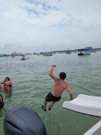 A young boy leaps off a boat into the water while others enjoy swimming nearby, capturing the excitement of a fun day on the water.