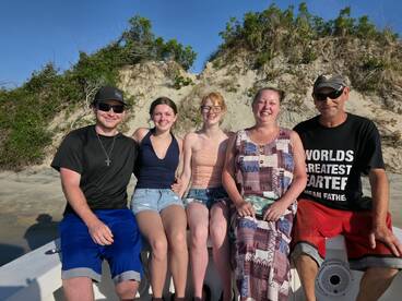 A cheerful group of family members poses together on a boat with a sandy beach backdrop, capturing a joyful moment during their seaside adventure.