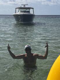 A man joyfully poses in shallow water with a boat anchored nearby, capturing the essence of fun and relaxation on the water.