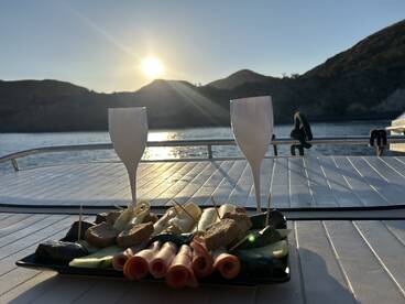 A beautifully arranged charcuterie platter with cocktails set against a stunning sunset backdrop on the water, perfect for a relaxing evening on a boat.