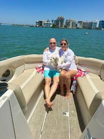 A couple enjoys a sunny day on their boat with a dog, showcasing a perfect moment of relaxation on the water against a vibrant city backdrop.