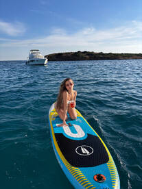 A young woman enjoys paddleboarding with a refreshing cocktail in hand, with a boat anchored in the background — a perfect day on the water.