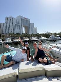 A cheerful group of friends enjoys their time aboard a boat in a marina, celebrating with smiles and waves against a backdrop of luxury yachts and a hotel.