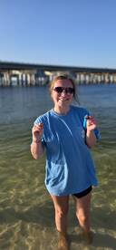 A joyful moment captured as a woman stands in shallow water, showcasing her catch while enjoying a sunny day by the pier — perfect for memorable beach outings.