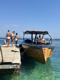 A lively group enjoys a sunny day by the water, with a stylish boat docked nearby — perfect for beach parties and summer fun.