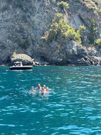 A joyful group swims in the crystal-clear waters near a rocky coastline, with a boat anchored nearby — perfect for family adventures and summer fun.