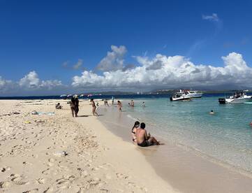 A lively beach scene with groups of people enjoying the sun and water, while boats are anchored nearby — perfect for a day of fun and relaxation on the shore.