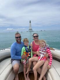 A joyful family poses on their boat with a lighthouse in the background, capturing a memorable day on the water with kids enjoying the adventure.