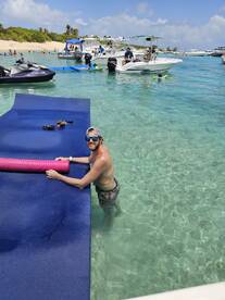 A man enjoys a sunny day at the beach, interacting with a floating mat while boats and fellow beachgoers create a lively atmosphere — perfect for summer fun and water activities.