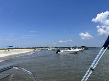 A vibrant scene at a sandbar with multiple boats anchored nearby, where people enjoy the beach and water activities under a clear blue sky.