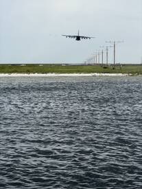 A distant view of an aircraft approaching for landing over a body of water, with power lines and vehicles visible in the background.