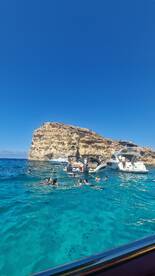 A lively scene of guests enjoying a swim in crystal-clear waters near a rocky coastline, with boats anchored nearby — perfect for snorkeling and beach fun.