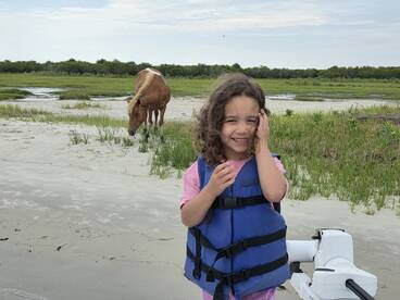 A joyful child in a life jacket smiles while a wild horse grazes nearby, showcasing a delightful moment of human-animal interaction in a natural setting.