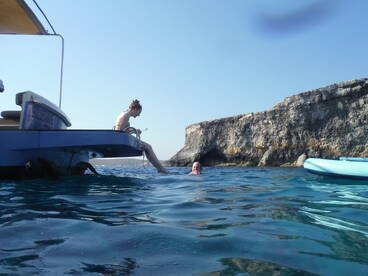 A woman sits on the edge of a boat while another person enjoys a swim in clear waters near a rocky coastline, capturing the essence of a fun day on the water.