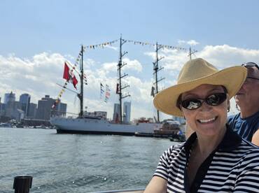 A joyful moment captured on a boat with a smiling woman in a straw hat, showcasing the vibrant city skyline and a tall ship in the background — perfect for memorable outings on the water.