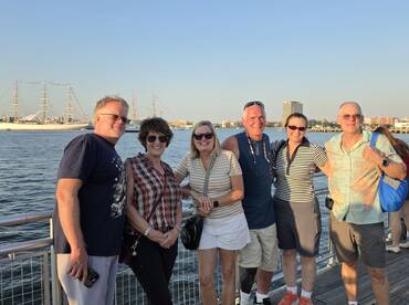 A cheerful group of friends poses together at a waterfront, enjoying a sunny day with a tall ship in the background — perfect for memorable outings and celebrations.