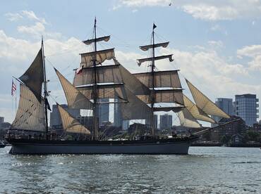 A majestic sailboat with billowing sails navigates the water, showcasing its classic design against a backdrop of a bustling city skyline.