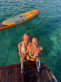 Two friends enjoy a refreshing swim and pose for a photo while holding onto the ladder of a boat, with a paddleboard nearby — a perfect moment of fun in the sun.