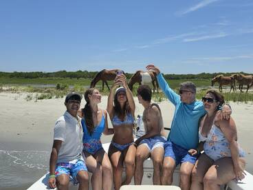 A lively group takes a selfie on a boat while wild horses graze on the beach in the background — a perfect blend of adventure and nature.