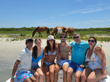 A joyful group poses on a boat with wild horses grazing in the background, capturing a memorable beach day filled with fun and adventure.