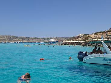 A vibrant scene at a popular beach with people swimming in crystal-clear turquoise waters, enjoying a sunny day with boats anchored nearby.