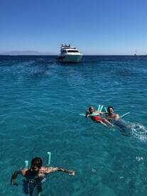 A lively group enjoys a sunny day swimming in crystal-clear waters, with a yacht anchored nearby — perfect for a fun day on the water.