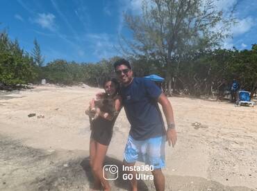 A joyful moment captured with a child and an adult holding a pig on a sandy beach, showcasing a unique human-animal interaction in a tropical setting.
