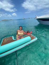 A joyful moment captured as a woman and a child enjoy swimming on an inflatable mat in crystal-clear waters, surrounded by boats — perfect for family fun on the beach.