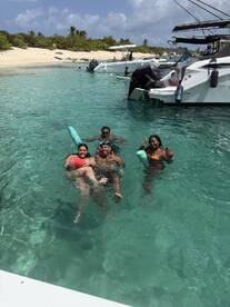 A lively group enjoys a fun day swimming in crystal-clear waters near a beach, with pontoon boats anchored nearby — perfect for a memorable day on the water.