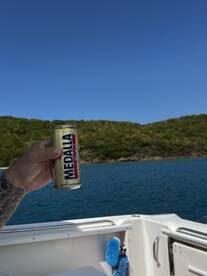 A hand holds a can of Medalla beer against a stunning backdrop of blue waters and greenery, capturing the essence of a relaxing day on the boat.