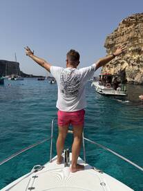 A man celebrates on the bow of a boat, enjoying the vibrant blue waters and scenic cliffs — a perfect moment for summer adventures on the sea.