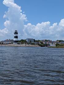 A picturesque view of a black and white lighthouse against a backdrop of fluffy clouds and coastal homes, perfect for capturing the charm of waterfront destinations.