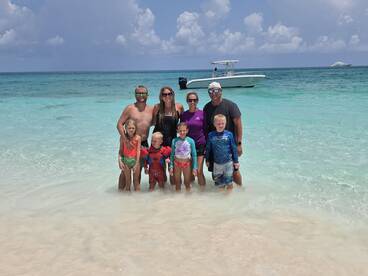 A joyful family poses together in shallow waters at the beach, with a boat anchored nearby — a perfect moment capturing fun and relaxation on a sunny day.