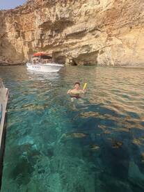 A swimmer enjoys the crystal-clear waters near a stunning cave, with a boat anchored nearby — perfect for snorkeling adventures and exploring coastal beauty.