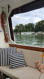 A cozy interior shot of a boat featuring striped cushions and a hand holding a snack, with a scenic view of the water and other boats in the background.