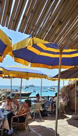 Guests enjoy a relaxing meal under colorful umbrellas at a seaside dining area, with boats visible in the background — a perfect spot for enjoying the coastal atmosphere.