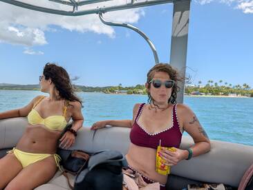 Two friends enjoy a sunny day on a boat, relaxing in their bikinis while taking in the beautiful waterfront scenery — perfect for a fun day out on the water.