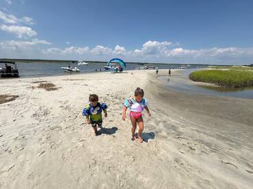 Two children explore a sandy beach area near a sandbar, surrounded by boats — a perfect family day on the water.