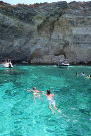 A couple enjoys a refreshing swim in crystal-clear waters, surrounded by stunning cliffs and boats — a perfect summer getaway.