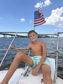 A young boy smiles while sitting on the bow of a boat, with an American flag waving in the background — a perfect moment capturing the joy of a day on the water.