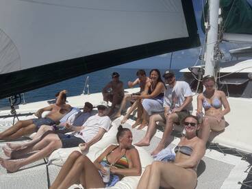 A lively group enjoys a day of sailing on a catamaran, relaxing on deck with drinks in hand — perfect for a fun day on the water.