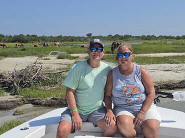 A couple enjoys a scenic boat ride with wild horses grazing in the background, capturing a perfect moment of nature and adventure.