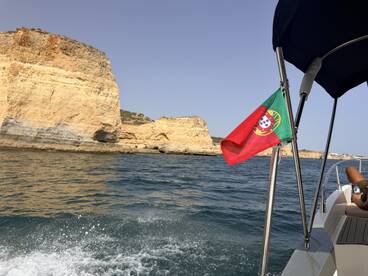 A scenic view of the rugged coastline of Portugal from a boat, showcasing the vibrant colors of the landscape and the Portuguese flag fluttering in the breeze.