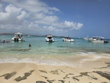 A vibrant scene at a sandbar with guests enjoying swimming and relaxing near boats in crystal-clear waters — a perfect day for beach lovers and water activities.