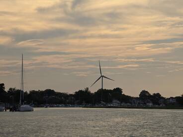 A serene sunset view featuring a sailboat and a wind turbine along the coastline, capturing the tranquil beauty of the waterfront.