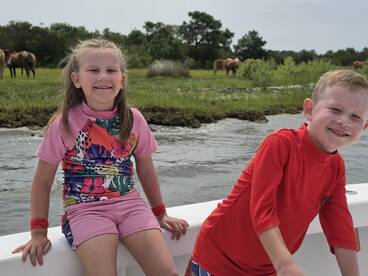 Two children enjoy a fun day on the boat, smiling as they take in the scenic view of wild horses grazing nearby — a perfect family adventure on the water.