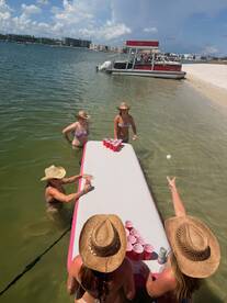 A lively group enjoys a fun beach day playing beer pong in the water, with a pontoon boat anchored nearby — perfect for summer celebrations and group activities.