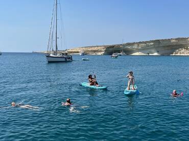 A lively group enjoys paddleboarding and swimming in crystal-clear waters, with a sailboat anchored nearby, creating the perfect backdrop for a fun day on the water.