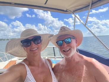 A cheerful couple takes a selfie on a boat under a bright blue sky, enjoying a sunny day on the water.