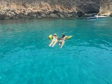 Two friends enjoy a relaxing day floating in crystal-clear waters, with a boat anchored nearby — a perfect summer getaway.